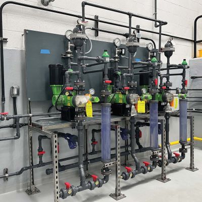 A water treatment or chemical dosing system featuring multiple green pumps, gray PVC piping, flow meters, and gauges, all mounted on a metal frame against a white and gray industrial wall.