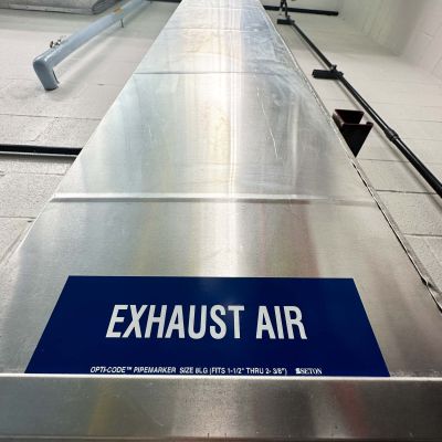 A low-angle view of a large, shiny, rectangular metal HVAC duct with a blue label reading 'EXHAUST AIR' running high across a white industrial ceiling.