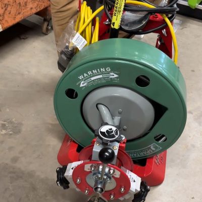 A close-up of a Spartan drain cleaning machine with a green drum and red frame, showing the coiled yellow cable and an attached root-cutting blade tool on the front.