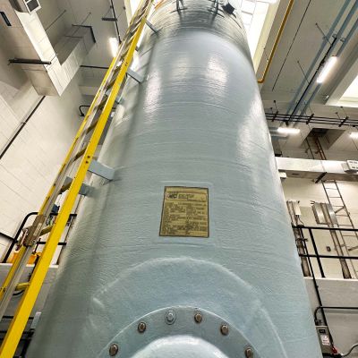 A low-angle shot of a tall, light blue industrial tank or pressure vessel with a yellow access ladder mounted on the side, stretching toward a skylight in the ceiling.