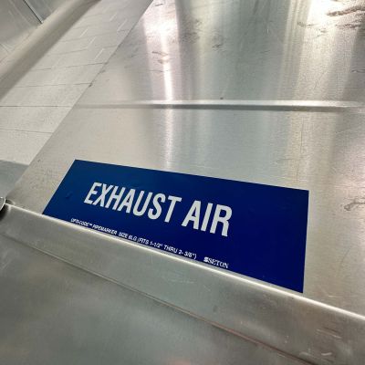 A close-up of a blue label with white text reading 'EXHAUST AIR' applied to a large, reflective silver metal HVAC duct in a commercial building.