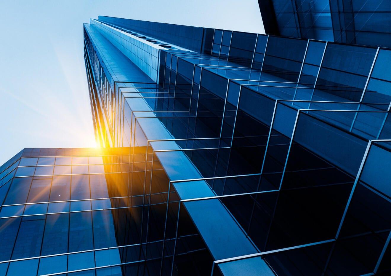 A dramatic, low-angle shot of a modern glass skyscraper reaching into a bright sky, with a burst of orange sunlight reflecting off the blue facade