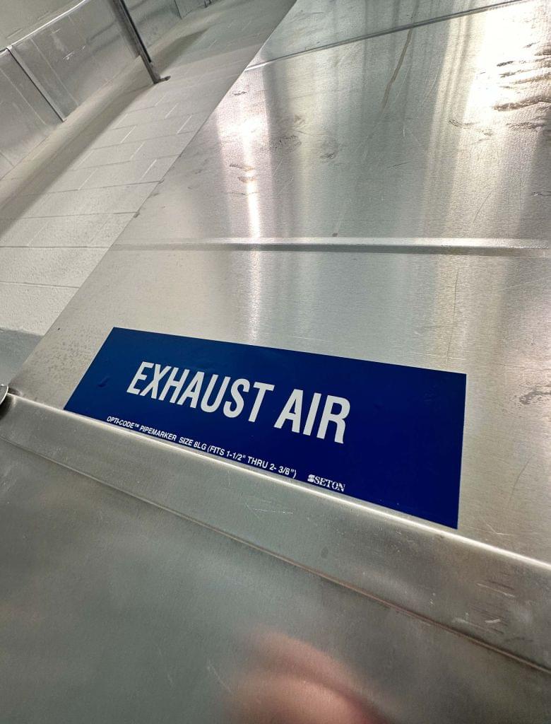A close-up of a blue label with white text reading 'EXHAUST AIR' applied to a large, reflective silver metal HVAC duct in a commercial building.